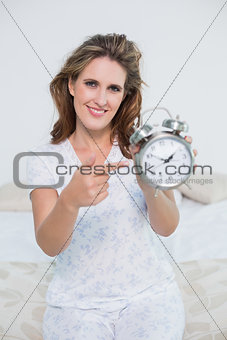 Smiling woman sitting on bed pointing at alarm clock