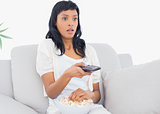 Shocked black haired woman in white clothes watching tv while eating popcorn