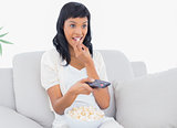 Focused black haired woman in white clothes watching tv while eating popcorn