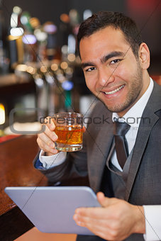 Cheerful businessman working on his tablet while having a whiskey