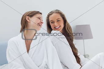 Happy friends wearing bathrobe sitting on bed