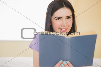 Cheerful girl looking at camera and lying on a bed reading a book