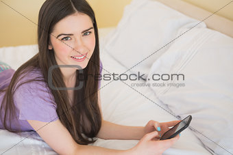 Happy girl looking at camera and lying on a bed using a mobile phone