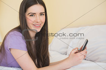 Cheerful girl looking at camera and lying on a bed using a mobile phone