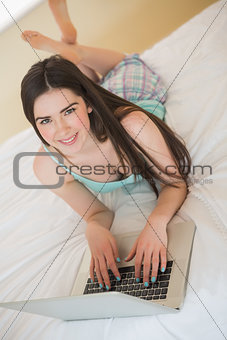 Brunette girl looking at camera and lying on a bed using a laptop
