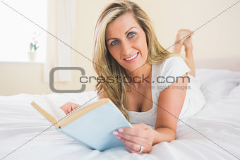 Joyful woman looking at camera reading a book lying on her bed