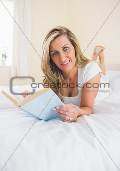 Pleased woman looking at camera reading a book lying on her bed