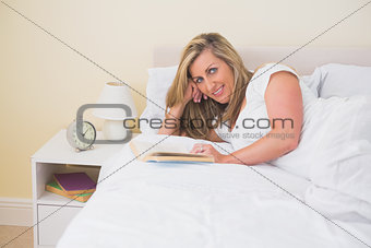 Content woman looking at camera reading a book lying on her bed