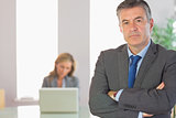 Unsmiling businessman looking at camera crossed arms with a businesswoman working on background