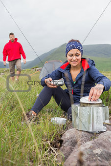 Woman cooking outside on camping trip with boyfriend walking
