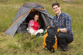 Smiling man packing backpack while girlfriend sits in tent