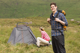 Smiling man carrying backpack while girlfriend is pitching tent