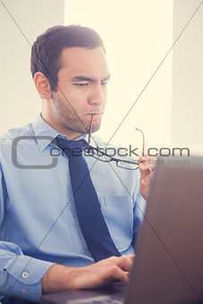 Focused man biting his eyeglasses and using a laptop