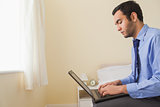 Unsmiling man using a laptop sitting on a bed