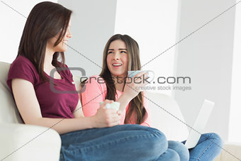 Two laughing girls sitting on a sofa enjoying a beverage and holding a laptop
