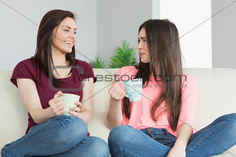 Two girls sitting on a sofa looking each other and drinking a beverage