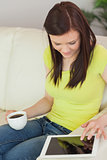 Smiling girl sitting on a sofa using a tablet pc and holding a cup of coffee