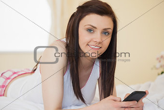 Smiling girl looking at camera using a mobile phone lying on a bed