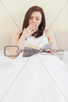 Yawning girl reading a book lying on a bed