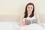 Smiling girl looking at camera and reading a book lying on a bed
