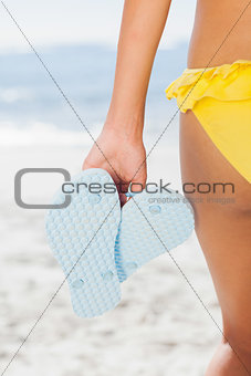 Woman in yellow bikini holding flip flops rear view