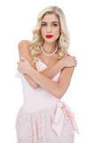 Severe blonde model in pink dress posing holding her shoulders and looking at camera