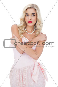 Severe blonde model in pink dress posing holding her shoulders and looking at camera