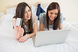Smiling friends using a laptop to shop online lying on bed