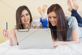 Cheerful friends using a laptop to shop online lying on bed