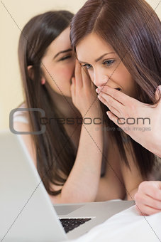 Pretty girl telling a secret to her friend in front of laptop
