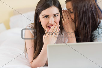 Happy girl telling a secret to her friend in front of laptop