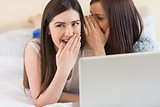 Smiling girl telling a secret to her friend in front of laptop