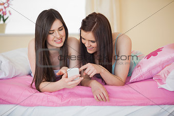 Smiling friends in pajamas looking at smartphone on bed