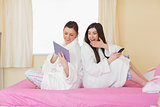 Two friends wearing bathrobes sitting back to back looking at tablet