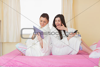 Two friends wearing bathrobes sitting back to back looking at tablet