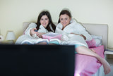 Two smiling friends wearing bathrobes watching a movie on bed