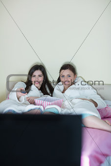 Two smiling friends wearing bathrobes watching tv on bed