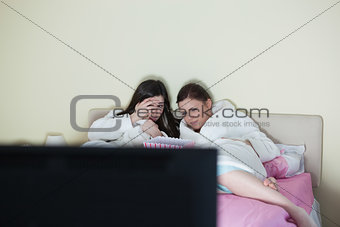 Two friends wearing bathrobes watching a scary movie on bed
