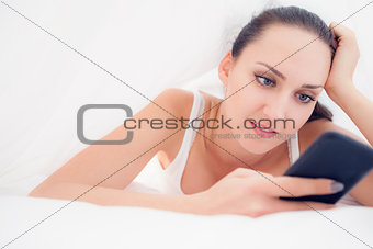 Happy brunette lying under the sheets using her smartphone