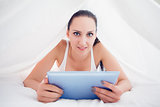 Happy brunette lying under the sheets holding her tablet pc