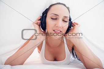 Happy brunette lying under the sheets listening to music