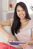 Pretty asian girl using her tablet on the couch smiling at camera
