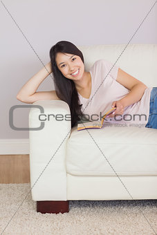 Content asian girl lying on the sofa reading book smiling at camera