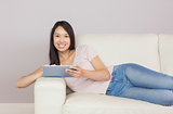 Pretty asian girl lying on the sofa using digital tablet smiling at camera