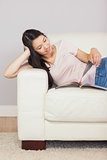 Cheerful asian girl lying on the couch reading a magazine
