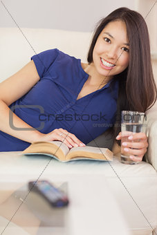 Pretty asian girl lying on the sofa reading a novel and drinking water