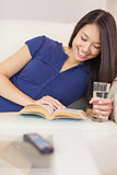 Smiling asian girl lying on the sofa reading a novel and drinking water