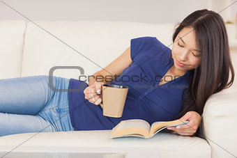 Beautiful asian girl lying on the sofa reading a novel and drinking hot beverage