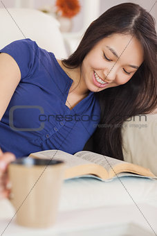 Relaxed asian girl lying on the sofa reading a novel and drinking hot beverage