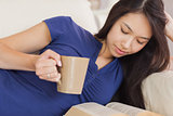 Attractive asian girl lying on the sofa reading a novel and drinking hot beverage